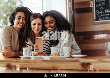 Tre donne seduti in un ristorante guardando al telefono cellulare e sorridente. Amici seduti in un caffè con caffè e snack sulla tabella guardando un mo Foto Stock