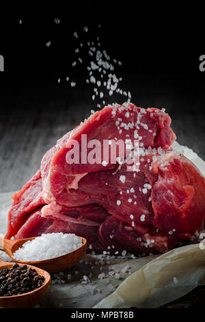 Un pezzo di carne cosparso con ampie sale di mare su di un tavolo di legno. Preparazione di carne per bistecche. Foto Stock