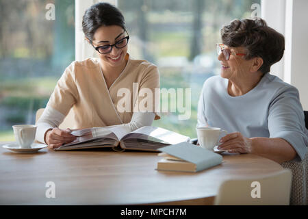 Felice donna senior di trascorrere del tempo insieme con una gara caregiver seduti a un tavolo e guardando album di foto di famiglia contro sfondo sfocato di w Foto Stock