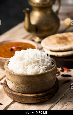 Primo piano di una ciotola con un korma di pollo al curry, una ciotola di riso cotto e alcuni chapatti su un bianco tavolo in legno con una teiera dorato nella parte posteriore Foto Stock