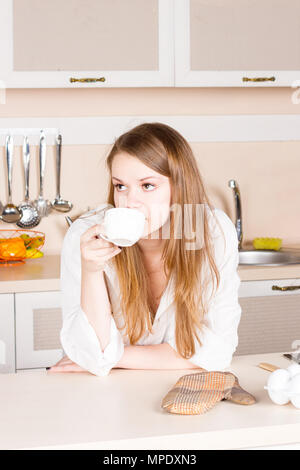 Ragazza in un bianco uomini shirt con lunghi capelli fluenti è bere il tè in cucina i gomiti sul tavolo al mattino. Inquadratura verticale Foto Stock