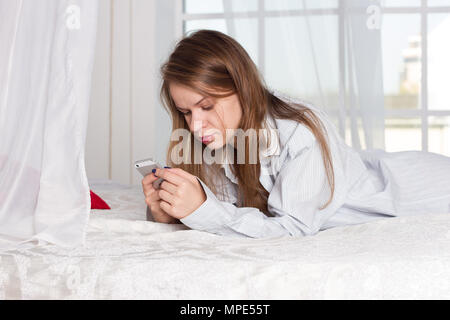 La ragazza si trova in un uomo shirt sul letto con uno smartphone nelle mani di Foto Stock