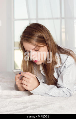 La ragazza si trova in un uomo shirt sul letto con uno smartphone nelle mani di Foto Stock