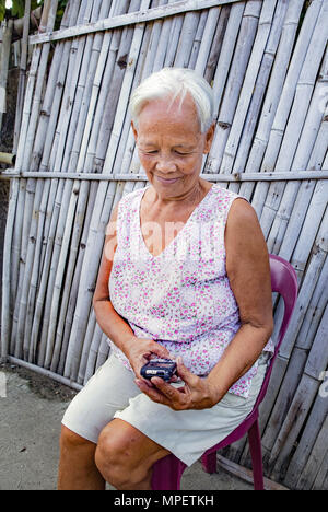 Senior, anziani bianco-dai capelli asiatici filippino donna seduta all'aperto provando a utilizzare il suo smartphone. Filippine. Foto Stock