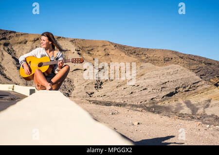 Scattante e uno stile di vita alternativo concept per il biondo modello caucasico sorridente outdoor con le montagne sullo sfondo e la riproduzione di una vecchia chitarra acustica Foto Stock