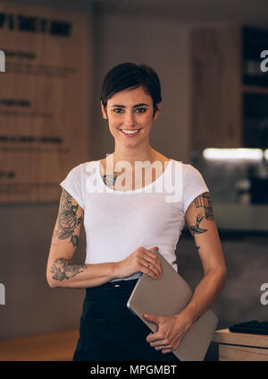 Ritratto di giovane donna in piedi in un ristorante in possesso di un computer portatile. Femmina caucasica con tatuaggi guardando la fotocamera e sorridente in un cafe'. Foto Stock