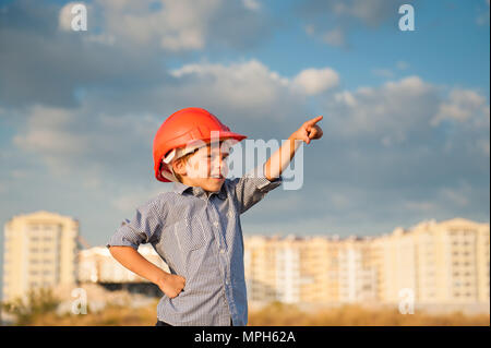 Carino piccolo ragazzo in arancione casco punti con il suo dito in direzione piedi su sfondo di edifici di nuova costruzione e di tramonto cielo molto nuvoloso Foto Stock
