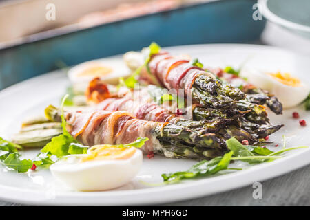 Asparagi freschi avvolto in pancetta su una piastra bianca con foglie di rucola pomodori e uova. Foto Stock
