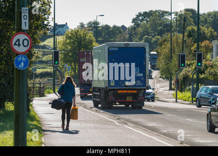 Una donna cammina lungo il marciapiede accanto ai veicoli che viaggiano lungo il bypass di Arundel, la A27 road, in Arundel, West Sussex, Regno Unito. Foto Stock