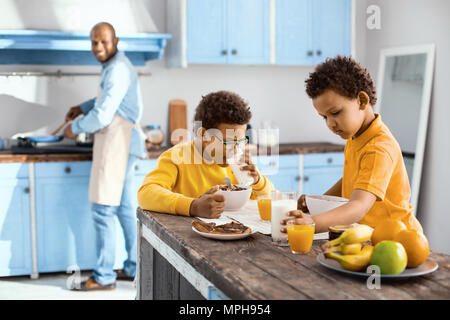 Piacevoli i bambini a mangiare la prima colazione durante il loro padre per la cottura Foto Stock