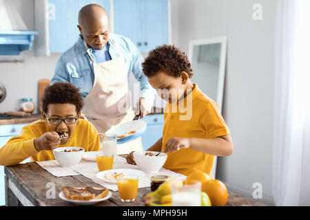 Simpatici ragazzi piccoli di mangiare la prima colazione durante il loro padre frittata di cottura Foto Stock