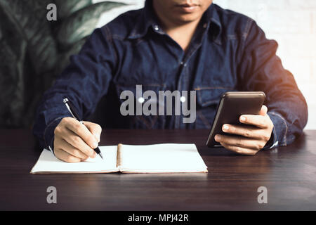 Imprenditore iscritto su notebook e guardando il telefono sul tavolo di legno Foto Stock