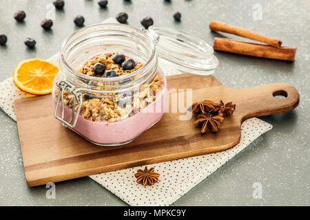 Rosa, yogurt alla frutta con granola e mirtilli in un recipiente su un tagliere di legno e cannella spice sul grigio di un contatore di cucina Foto Stock