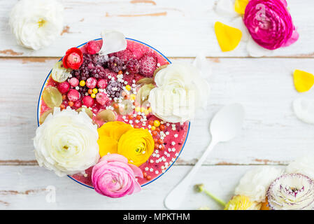 La colazione Detox frullato rosa da banana e frutti di bosco nella ciotola, guarnita con frutti di bosco surgelati e ranunculus fiori, luce sullo sfondo di legno, Foto Stock