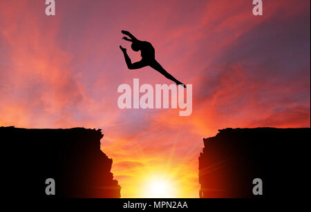 Silhouette di una ragazza ballerino in un cervo split leap su scogli pericolosi con drammatico tramonto o l'alba di sfondo e copiare lo spazio. Concetto di fede, conq Foto Stock