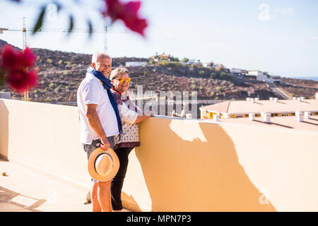 Coppia di anziani sulla terrazza a lui con pantaloncini e maglietta e hat ella con camicia e pantaloni guardare e sorridere all'aperto sulla terrazza con sole estivo Foto Stock