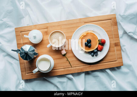 Vista in pianta di una prima colazione a letto con pane appena sfornato, frittelle e caffè sul vassoio in legno Foto Stock