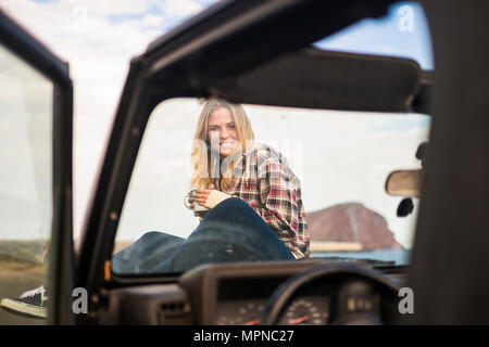 La libertà e il concetto di vacanza per la bella bionda caucasian giovane donna seduta sul naso di un fuoristrada nero auto. La retroilluminazione e la luce del sole in wa Foto Stock