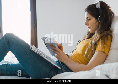 Giovane donna con il computer portatile e cuffie giacente sul letto a scrivere qualcosa di Foto Stock