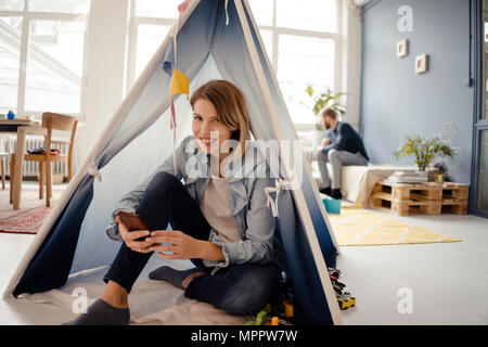 La donna utilizza lo smartphone in una tenda giocattolo, marito seduto in background Foto Stock