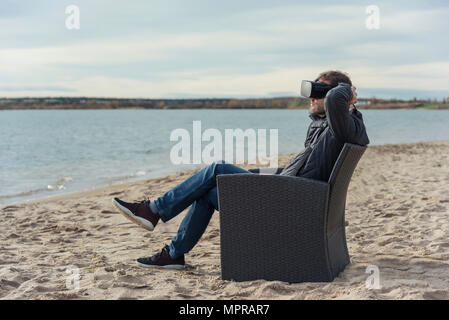 Uomo seduto in poltrona sulla spiaggia indossando occhiali VR Foto Stock