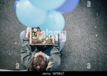 Donna con il presente in una scatola di cartone e palloncini blu seduto a terra Foto Stock