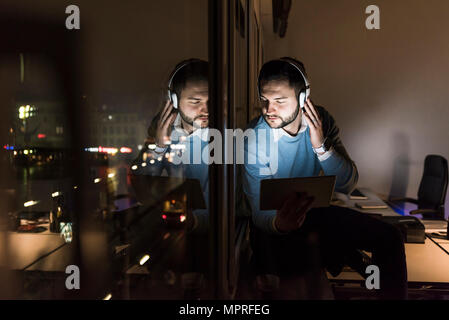 Imprenditore seduto sul davanzale in ufficio in serata ascoltando musica con le cuffie Foto Stock