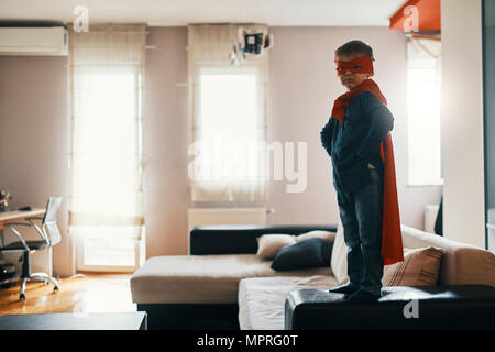 Little Boy vestito come un supereroe in piedi sul tavolo da caffè a casa Foto Stock