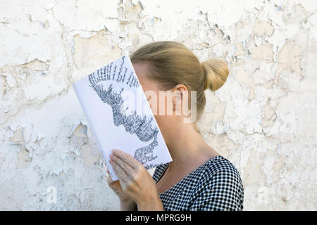 Donna che ricopre la faccia con il libro e la lettura di poesie nella parte anteriore della parete Foto Stock