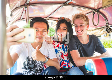 Thailandia, Bangkok, tre amici in sella tuk tuk tenendo selfie con lo smartphone Foto Stock