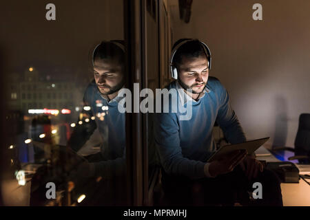 Imprenditore seduto sul davanzale in carica durante la notte utilizzando tablet e cuffie Foto Stock