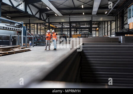 Due uomini che indossano copricapi rigidi e giubbotti di sicurezza parlando al negozio della fabbrica piano Foto Stock