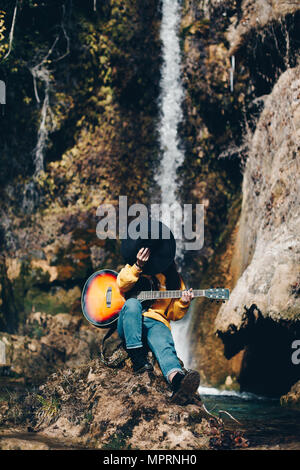 Spagna, Lleida, giovane donna con la chitarra seduto sulla roccia di fronte a cascata che ricopre la faccia con hat Foto Stock