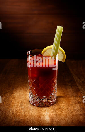 Riflettori puntati su un cristallo di bicchieri pieni di Bloody Mary cocktail, con un sedano guarnire, girato su un antico tavolo di legno alto. Foto Stock