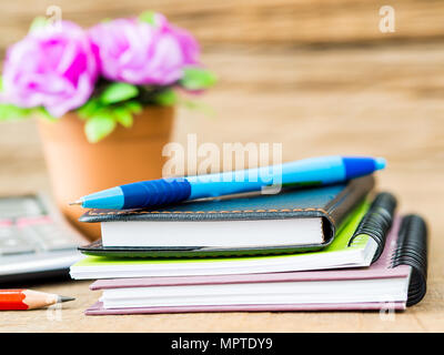 Accessori per ufficio tra cui nota libri, penna blu, calcolatrice e fiore su sfondo di legno. L'istruzione e la concezione di business. Foto Stock