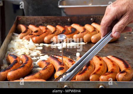 I vecchi mano maschio con pinze girando la salsiccia Hot Dogs, cipolle su una griglia esterna, grigliare venditore ambulante. Americana popolare venditore ambulante cuisi Foto Stock