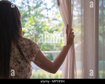 Vista posteriore di una giovane donna che mantiene le tende aperte a guardare attraverso la finestra. È disperata e triste donna concetto. Foto Stock