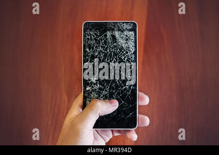 Smartphone rotto in mano azienda. guy sostiene il telefono con un display rotto vetro in mano sul background del pavimento in legno. Spaccatura su un vetro di colore nero Foto Stock