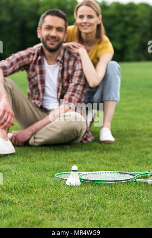 Volano e badminton racchette giacente su erba, felice coppia seduta dietro Foto Stock