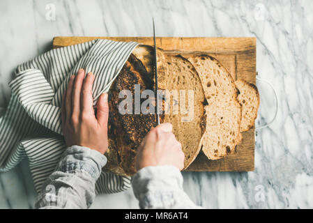 Flat-lay di mani femminili il taglio fresco cotto al forno del pane di pasta acida in pezzi su rustiche tagliere di legno su grigio chiaro sfondo marmo, vista dall'alto Foto Stock