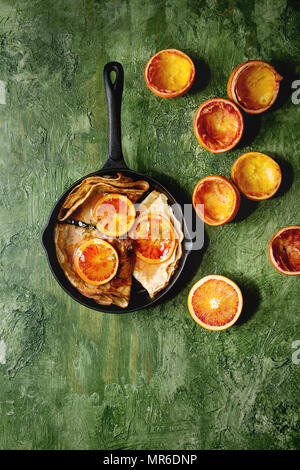 In casa Crepes Pancake servita in ghisa padella con sanguinose arance e sciroppo di rosmarino con fette di arance rosse di Sicilia su una texture verde sullo sfondo Foto Stock