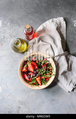 La barbabietola e insalata di fragole servite con foglie di rucola e i dadi sul vaso di ceramica con un panno e bottiglie di frutta ocet e olio di oliva su grigio backgr texture Foto Stock
