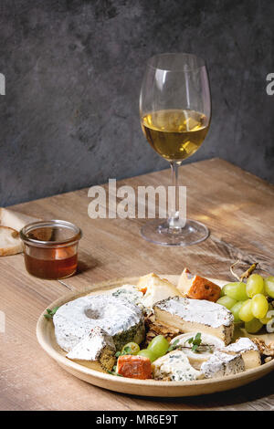 Il piatto di formaggi assortimento di formaggi francesi servite con miele, noci, pane e uva su piastra in ceramica e vetro di vino bianco su un tavolo di legno con Foto Stock