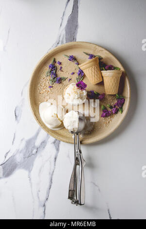 In casa di gelato alla vaniglia sfere servita con viola fiori commestibili e cucchiaio di metallo nella piastra in ceramica di colore bianco su sfondo marmo. Vista superiore, spazio. Foto Stock