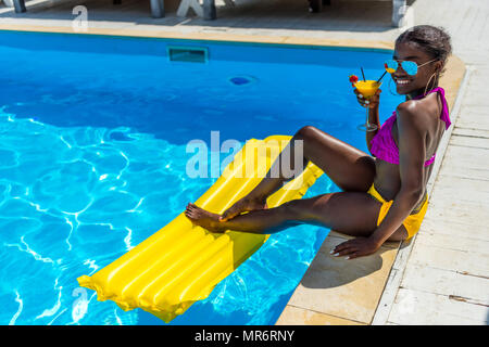 African American donna appoggiata con cocktail vicino alla piscina presso il resort Foto Stock