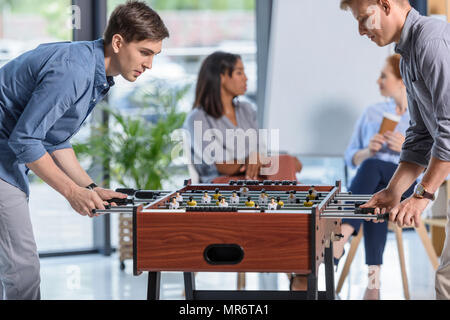Due imprenditori a giocare il calcio balilla mentre i giovani imprenditrici in chat durante la pausa in ufficio moderno Foto Stock