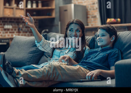 Piccolo figlio e il suo giovane madre parlare mentre si guarda la TV sul divano e mangiare popcorn Foto Stock