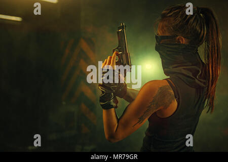 Pericoloso combattente donna con la pistola e sciarpa nel buio interiore con il fumo. Tatuaggio sul corpo. Foto Stock