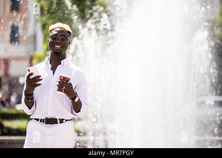 African American uomo seduto in città e utilizzando il suo telefono cellulare con le cuffie nel giorno di estate vicino alla fontana. Foto Stock