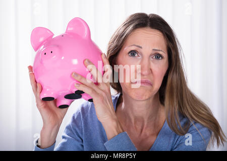 Close-up di donna infelice Holding Piggybank contro di persiane Foto Stock